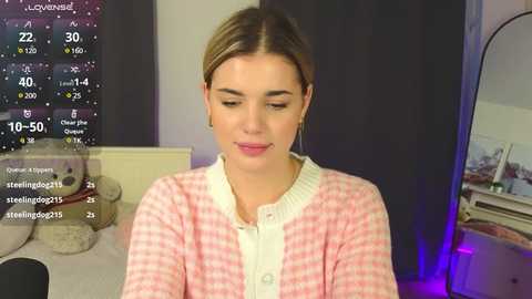 Media: Video of a young woman with fair skin, light brown hair, and blue eyes, wearing a pink checkered cardigan, sitting indoors.