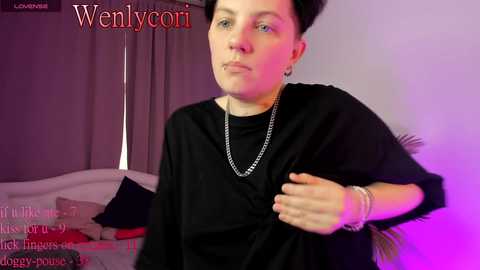 Media: Video of a fair-skinned woman with short dark hair, wearing a black robe, standing in a dimly lit room with purple lighting.