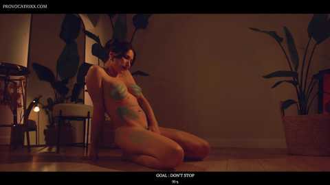 Media: A video of a nude woman with green body paint, kneeling on a wooden floor, surrounded by potted plants in a dimly lit room.