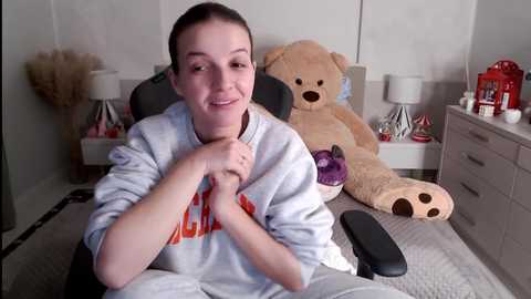 Media: Video of a smiling woman in a grey hoodie with a \"Ralph\" logo, sitting on a grey chair in a cozy bedroom with a teddy bear and a red Christmas tree.