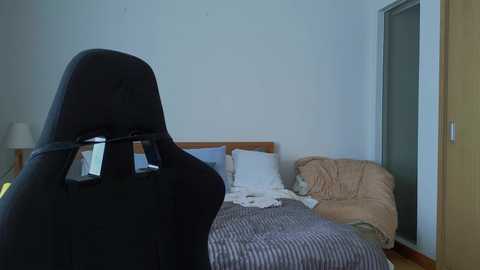 Media: Video of a messy bedroom with a black gaming chair facing the bed, which has white sheets, a beige blanket, and a striped duvet. The room's walls are light blue, and there's a door and a lamp in the background.