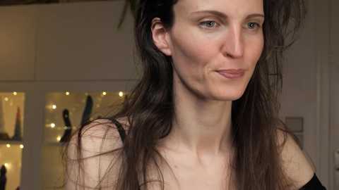 Media: Video of a fair-skinned woman with long, dark hair, wearing a black top, in a softly lit room with a glass cabinet displaying colorful items.