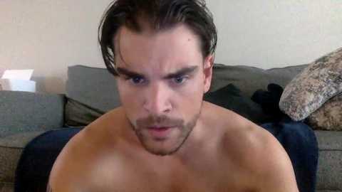 Media: Video of a shirtless man with short dark hair, fair skin, and a trimmed beard, looking serious. Background features a grey sofa with a plush pillow, beige walls, and a white object.