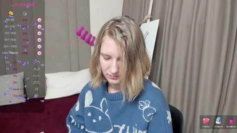 Media: Video of a young woman with shoulder-length blonde hair, wearing a blue sweater adorned with white bunny designs, sitting on a bed with beige curtains and a red blanket. A pink hair clip is visible.