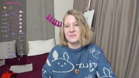 Media: Video of a young woman with shoulder-length blonde hair, wearing a blue sweater with cartoon rabbits and a purple spiral hair accessory, standing in a bedroom with a white bed and gray curtains.