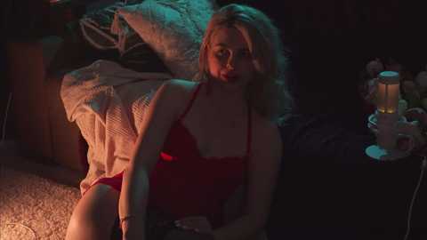 Media: Video of a young woman with blonde hair, fair skin, wearing a red lace bra and panties, sitting on a carpeted floor in dim lighting.