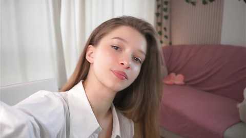 Media: Video of a young Caucasian woman with long brown hair, wearing a white blouse, smiling slightly, indoors with a pink couch and white curtains in the background.