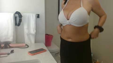 Media: Video of a woman in a white bra and black leggings, hands on hips, standing in a modern bathroom with a white countertop, towels, and pink trash can.