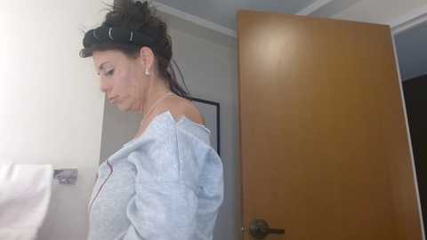 Media: Video of a woman with dark hair, wearing a black headband and light gray hoodie, standing in a bathroom with a wooden door and white towel on a rack.