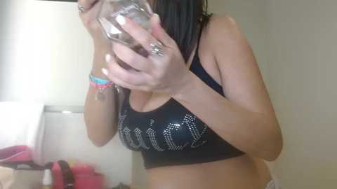 Media: Video of a woman with medium brown skin and straight, dark hair, wearing a black sports bra with silver studs, taking a selfie with a glass in front of her face, in a dimly lit room with a bed and clothing visible in the background.