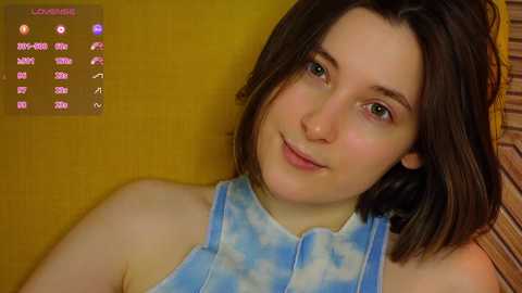 Media: A video of a young woman with fair skin, dark brown hair, and green eyes, wearing a blue tie-dye top, smiling against a mustard-yellow wall.