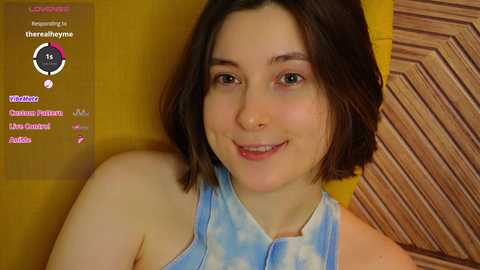 Media: Video of a young Caucasian woman with shoulder-length brown hair, wearing a light blue tie-dye top, smiling, against a wooden textured background.