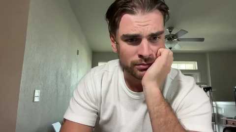 Media: Video of a man with light skin and short brown hair, wearing a white shirt, resting his chin on his hand, looking contemplative in a beige room with white walls and a ceiling fan.