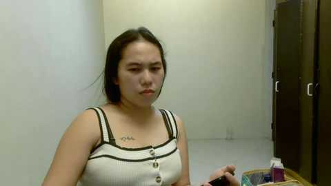 Media: Video of a young Asian woman with straight black hair, wearing a white tank top with black straps and buttons, standing in a small, plain room with beige walls and a dark wooden cabinet.