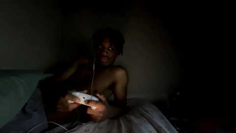 Media: A dimly lit video of a nude African-American woman with medium skin tone, sitting on a bed, holding a white game controller, with headphones on, in a dimly lit room.
