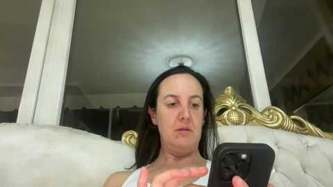 Media: A video of a woman with long dark hair, wearing a white tank top, sitting on a white tufted couch with a gold headboard, taking a selfie with a black phone.