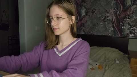 Media: Video of a young girl with straight, light brown hair, wearing glasses, a purple sweater, and sitting on a bed with a green blanket. The background features a dark, floral-patterned wall and scattered objects.