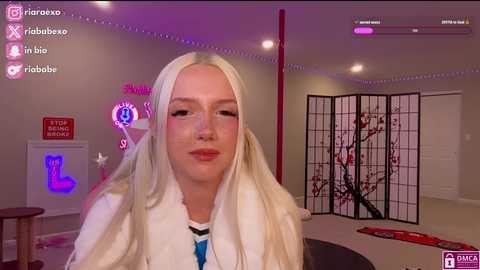 Media: A video of a young, fair-skinned woman with long, straight platinum blonde hair, wearing glasses and a white sweater, sitting in a dimly lit room with pink lighting, a black screen divider, and a red pole.