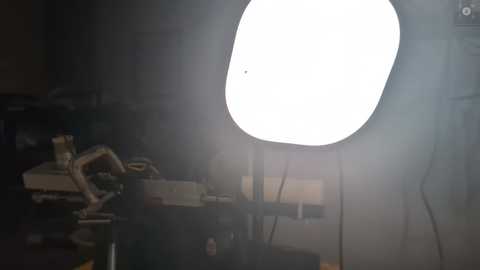 Media: Video of a dimly lit studio with a large, bright, white softbox light illuminating a blurred subject. The background is shadowy, with various studio equipment and objects barely visible.