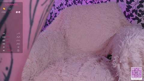 Media: Video of a fluffy white cat curled up on a soft, cream-colored cushion, surrounded by a pink and black floral wallpaper. The cat's fur is light and textured, contrasting with the plush cushion.
