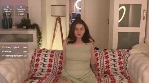 Media: A video of a young woman with fair skin and long dark hair, wearing a light green dress, sitting on a patterned couch in a cozy living room.