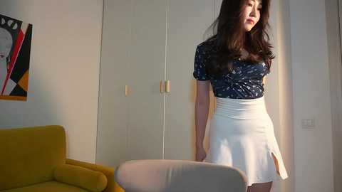 Media: Video of a young Asian woman with long dark hair, wearing a navy blue floral top and a white skirt with a slit, standing in a modern, minimalist living room with a mustard-yellow couch and abstract artwork on the wall.