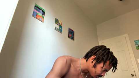 Media: Video of a shirtless, light-skinned man with dreadlocks, wearing a silver necklace, standing in a minimalist room with white walls, colorful abstract paintings, and a closed white door.