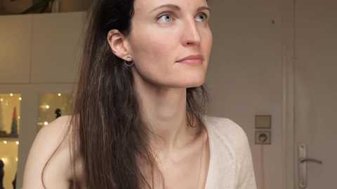 Media: Video of a fair-skinned woman with long, straight brown hair, wearing a white top, standing indoors with a soft, neutral background featuring a closed door and a wall-mounted light switch.