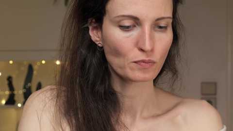 Media: Video of a fair-skinned woman with long, straight, dark brown hair, wearing small hoop earrings, in a neutral expression, indoors with blurred background.