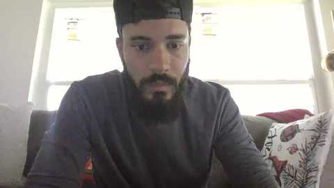 Media: Video of a bearded man with a black cap, wearing a gray long-sleeve shirt, seated on a couch with floral-patterned cushions, in a brightly lit room with white blinds.