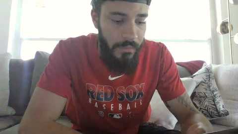 Media: A video of a bearded man in a Boston Red Sox baseball jersey, sitting on a couch with cushions. He appears focused, with a serious expression. The background features a white window and a gray couch.