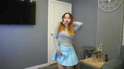 Media: Video of a young woman with straight, shoulder-length blonde hair, wearing a light blue crop top and mini skirt, posing in a modern living room with a flat-screen TV, a clock, and a glass table.