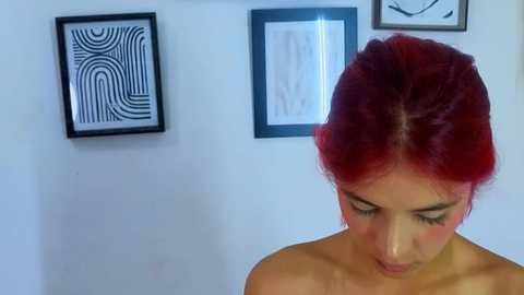 Media: Video of a topless woman with vibrant red hair styled in a high ponytail, looking down, set against a minimalist white wall with three black-framed abstract art pieces.