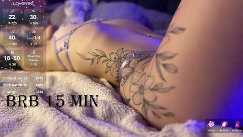 Media: A close-up video of a woman's lower back and arm, showcasing intricate floral tattoos and a delicate, light-colored bra. The background is blurred, featuring a soft, textured blanket and a digital timer display.