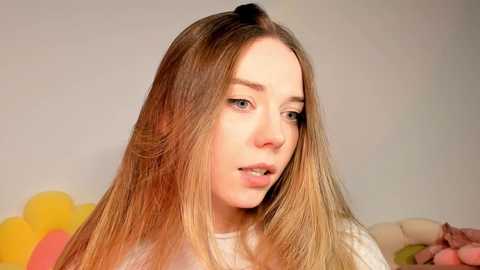 Media: Video of a young woman with long, light brown hair, pale skin, and blue eyes, wearing a white top, looking contemplative against a soft-focus, light-colored background.
