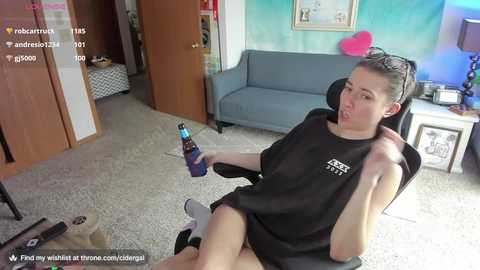 Media: Video of a woman with light skin and dark hair in a ponytail, wearing a black shirt and shorts, sitting in a gaming chair, holding a beer bottle, in a cozy living room with a blue sofa.