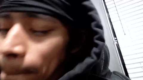 Media: Video of a man with a scruffy beard, wearing a black hooded jacket, blurred in the foreground, standing near a window with white blinds.