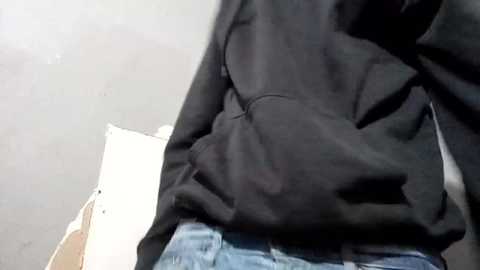 Media: Video of a person's torso wearing a black hoodie and blue jeans, hands in pockets. Background is a blurred, light-colored wall with a white chair on the left.