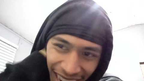 Media: A low-quality video of a man with light skin, wearing a black hoodie and a black face mask, smiling with a gap-toothed grin. The background shows a white ceiling and window blinds.