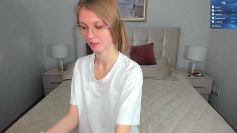 Media: Video of a slender, fair-skinned, young woman with straight, light brown hair, wearing glasses, and a white T-shirt. She stands in a modern, light-colored bedroom with a neatly made bed, two nightstands, and a TV screen displaying a streaming service interface.
