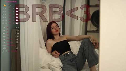 Video of a young Caucasian woman with long red hair, wearing a black strapless top and high-waisted jeans, lounging on a white bed with a large fan in the background, surrounded by a digital interface with \"BBB\" arrow and icons.