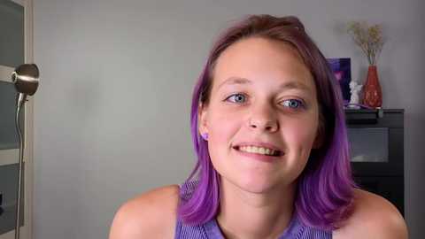 Media: Video of a cheerful young woman with shoulder-length purple hair, wearing purple earrings, a blue sleeveless top, smiling, indoors against a grey wall with a black cabinet and vase.