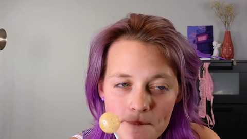 Media: Video of a woman with shoulder-length, purple hair, wearing a pink and white striped top, sucking on a yellow lollipop, in a modern room with purple decor.