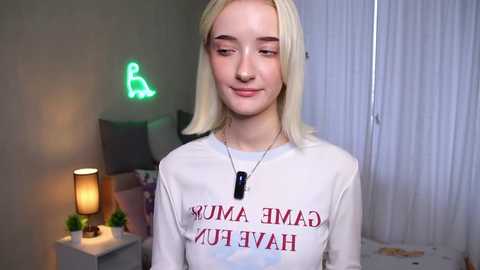 Media: Video of a young woman with straight platinum blonde hair, wearing a white \"HAIMAN JUMA\" t-shirt and a dog tag necklace. Background includes a bed, a green neon sign, and a lamp.