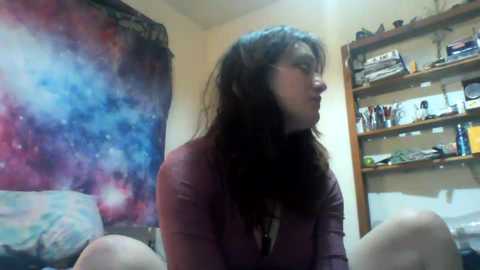 Media: A video of a woman with long, dark hair and a maroon cardigan, standing in a dimly lit room with a colorful starry wall and a cluttered bookshelf.
