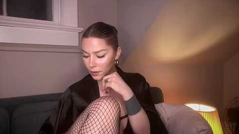 Media: Video of a woman with dark hair tied back, wearing a black blazer, fishnet stockings, and a grey wristwatch, sitting on a dark sofa. A soft-lit lamp and a window with white trim are in the background.