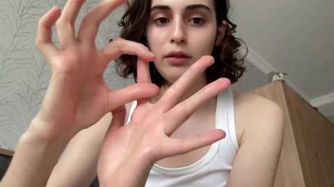 Media: Video of a young person with light skin, curly brown hair, and wearing a white tank top, hands raised in a gesture of \"OK.\" Background features light-colored, textured wallpaper and a wooden cabinet.