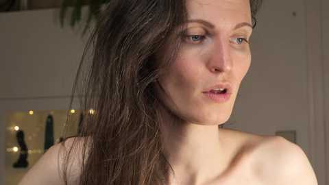 Media: A video of a woman with long, wavy brown hair, fair skin, and blue eyes, partially seen, in a softly lit room.