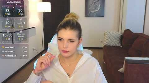 Media: Video of a young Caucasian woman with blonde hair tied in a bun, wearing a white hoodie, sitting on a brown couch in a modern living room. Weather app on the wall displays rain.