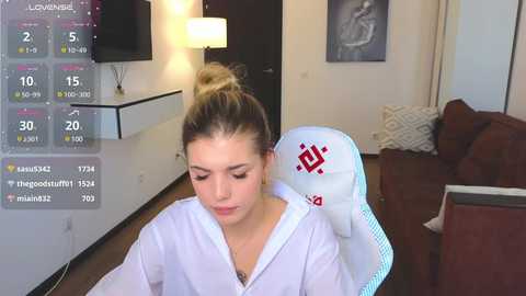 Media: Video of a young blonde woman in a white robe, seated in a white chair, watching a TV with a weather forecast displayed. Background features a brown sofa, white wall, and a lamp.